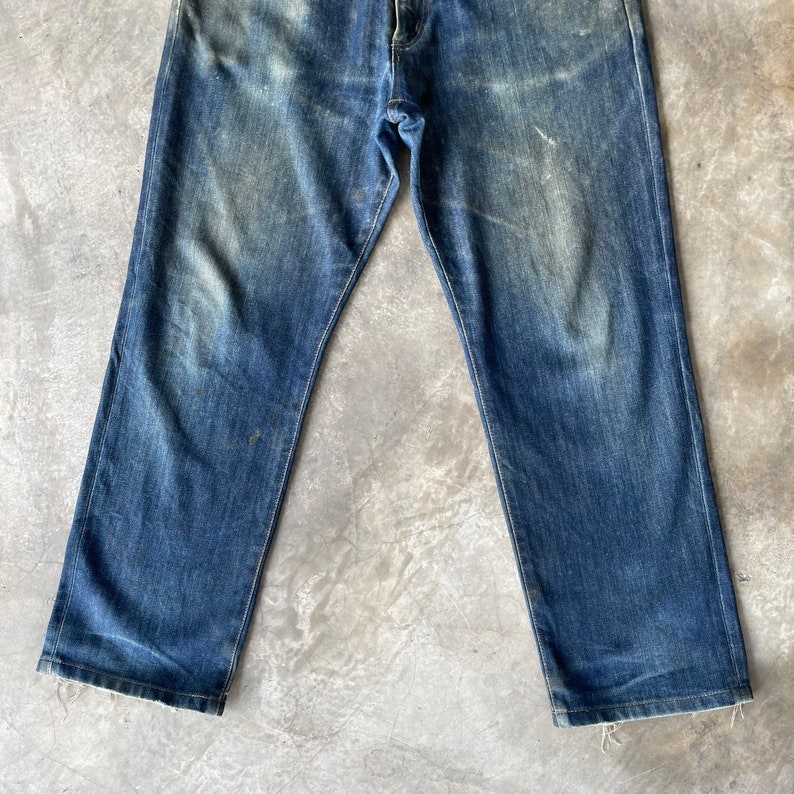 Puede incluir: Vaqueros de mezclilla azul descolorido con corte recto. Los vaqueros muestran signos de desgaste con parches m&aacute;s claros y sutiles desgastes. El dise&ntilde;o cl&aacute;sico de cinco bolsillos es visible, con costuras blancas.