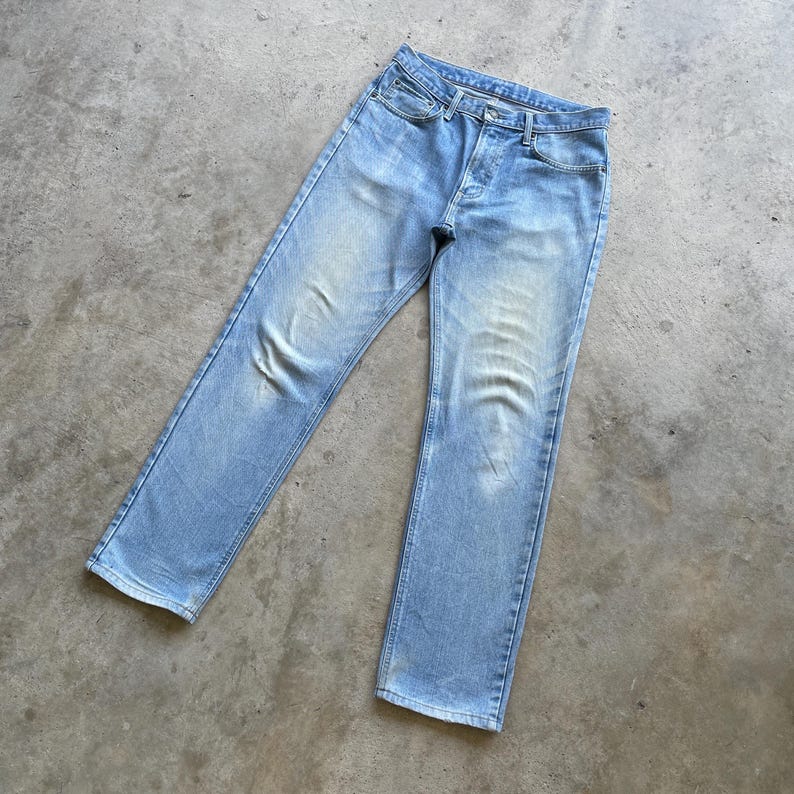 May include: Light blue denim jeans with a classic five-pocket design. The jeans have a button fly and a faded wash, giving them a vintage look. The jeans are laid flat on a concrete surface.