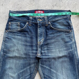 May include: Close-up of blue denim jeans with a faded wash. The jeans have a button closure and classic five-pocket design. A measuring tape shows a waist measurement of approximately 48 cm. The brand name "EDWIN" is visible on a red tag.