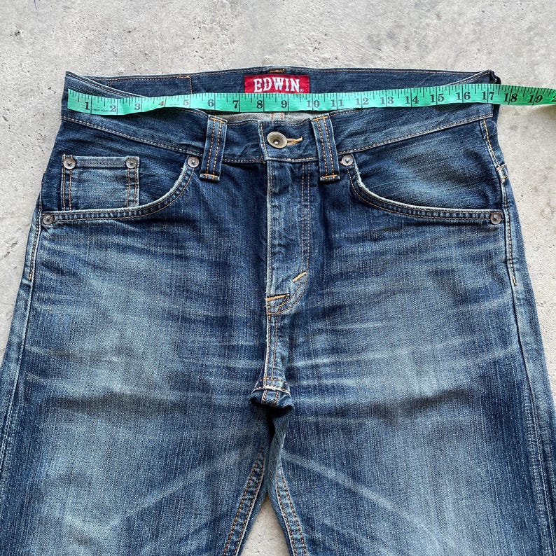 May include: Close-up of blue denim jeans with a faded wash. The jeans have a button closure and classic five-pocket design. A measuring tape shows a waist measurement of approximately 48 cm. The brand name "EDWIN" is visible on a red tag.
