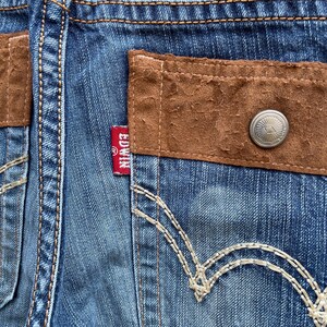 Puede incluir: Primer plano de vaqueros de mezclilla azul con detalles de bolsillo de gamuza marr&oacute;n. Los vaqueros presentan un bot&oacute;n plateado y la etiqueta roja de la marca Edwin. Las costuras blancas acent&uacute;an los bolsillos y las costuras.