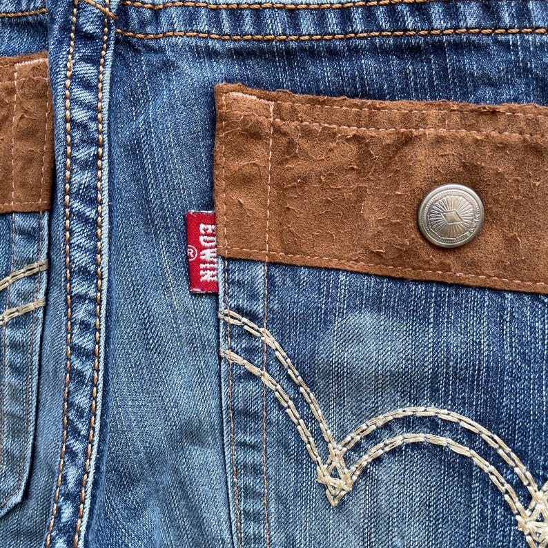 Puede incluir: Primer plano de vaqueros de mezclilla azul con detalles de bolsillo de gamuza marr&oacute;n. Los vaqueros presentan un bot&oacute;n plateado y la etiqueta roja de la marca Edwin. Las costuras blancas acent&uacute;an los bolsillos y las costuras.
