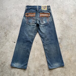 May include: Blue denim jeans with brown suede pocket accents. The jeans have a faded wash and a straight leg cut. The back pockets feature decorative stitching and a small red tag. A small patch is visible on the back waistband.