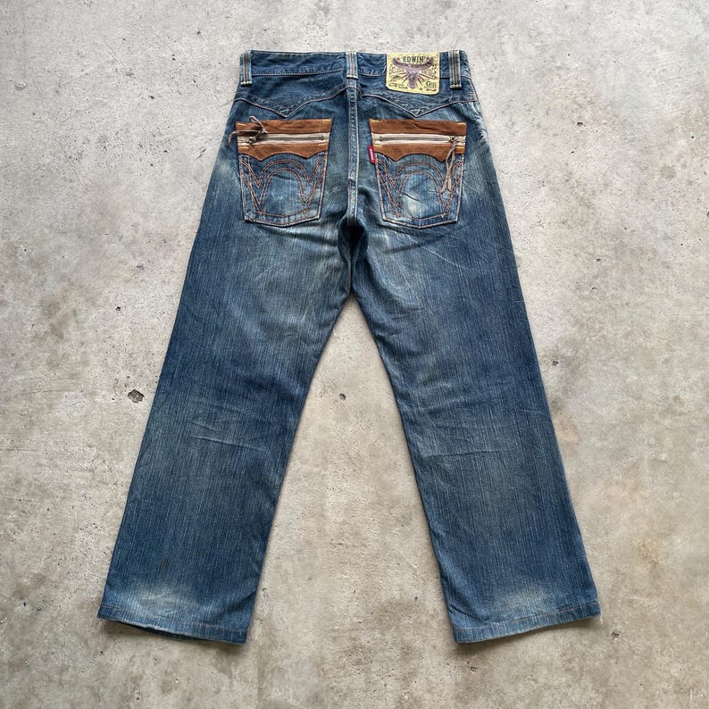 May include: Blue denim jeans with brown suede pocket accents. The jeans have a faded wash and a straight leg cut. The back pockets feature decorative stitching and a small red tag. A small patch is visible on the back waistband.
