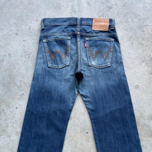 May include: Blue denim jeans with a classic five-pocket design. The back pockets feature a distinctive stitched pattern. A brown leather patch with the brand name is visible at the waistband. The jeans have a faded wash and a straight leg.