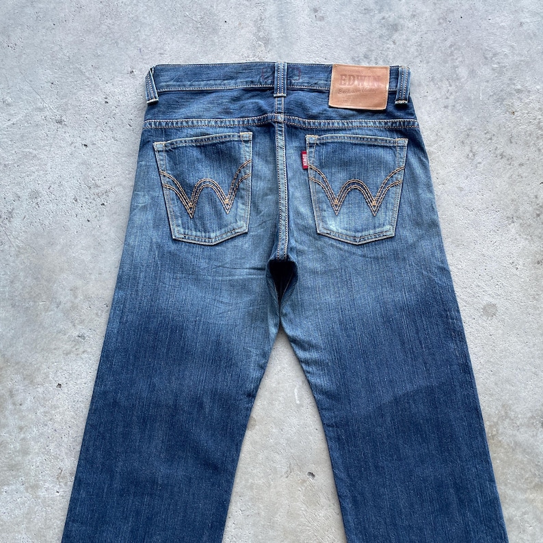May include: Blue denim jeans with a classic five-pocket design. The back pockets feature a distinctive stitched pattern. A brown leather patch with the brand name is visible at the waistband. The jeans have a faded wash and a straight leg.