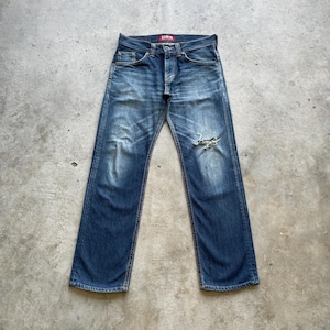 May include: Blue denim jeans with a distressed appearance, showing a faded wash and a ripped knee. The jeans have a classic five-pocket design and a red Edwin label. The jeans are laid flat on a concrete surface.