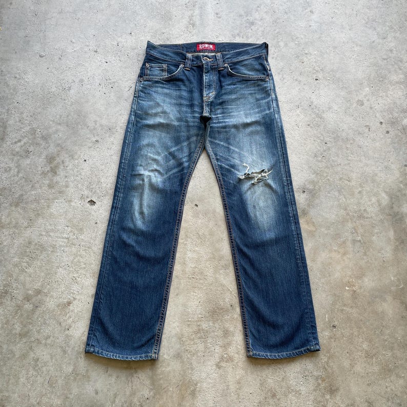May include: Blue denim jeans with a distressed appearance, showing a faded wash and a ripped knee. The jeans have a classic five-pocket design and a red Edwin label. The jeans are laid flat on a concrete surface.