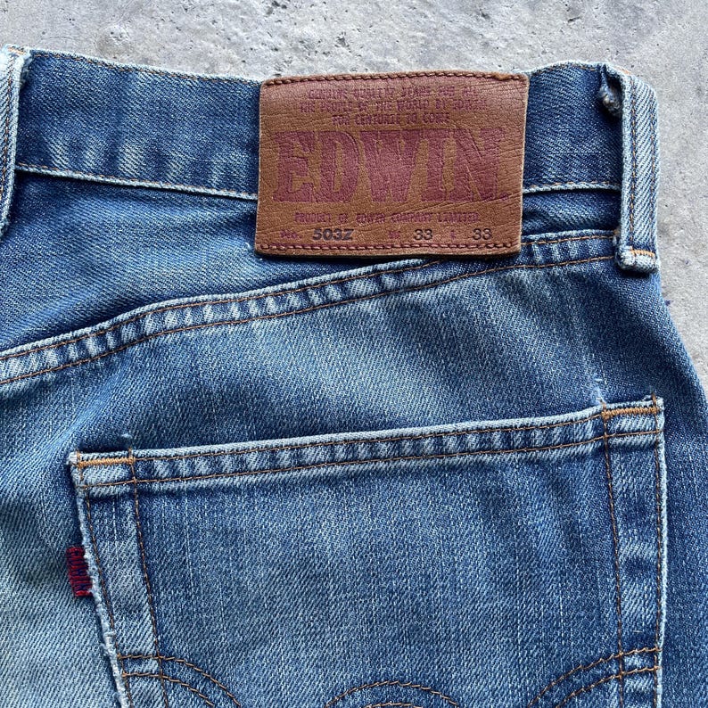 Pu&ograve; includere: Primo piano di jeans blu in denim con toppa in pelle marrone. La toppa presenta il marchio "EDWIN" in grassetto. I jeans hanno un classico design a cinque tasche con un'etichetta rossa sulla tasca posteriore. Misura della vita 84 cm.