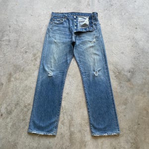 May include: Faded blue denim jeans with a button fly and distressed details. The jeans have a classic five-pocket design and a straight leg fit. The denim has a worn appearance with visible fading and some tears.