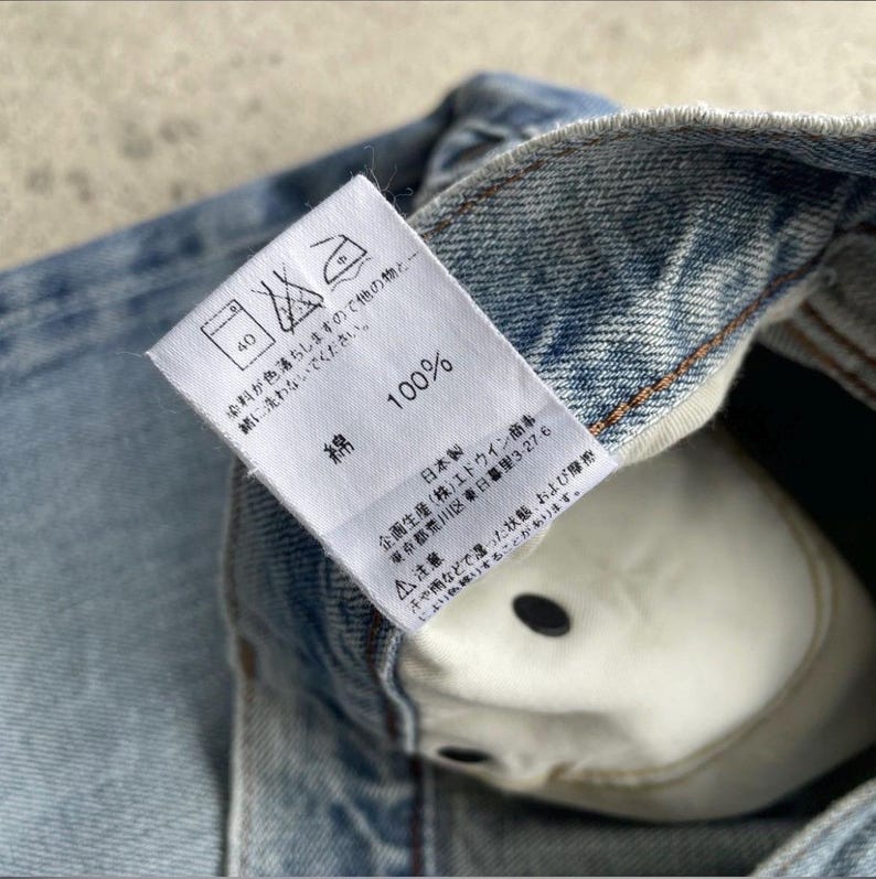May include: A close-up shot of light blue denim fabric with a white label. The label shows washing instructions, the number 40, and the text "100% 綿". The garment's interior reveals a white lining and a black button.