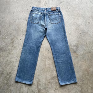 May include: Light blue denim jeans with a classic five-pocket design. The jeans have a straight leg cut and a worn appearance. The back pockets feature a distinctive stitched design. A brown leather patch is visible at the waistband.