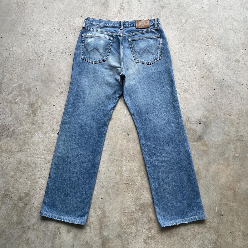 May include: Light blue denim jeans with a classic five-pocket design. The jeans have a straight leg cut and a worn appearance. The back pockets feature a distinctive stitched design. A brown leather patch is visible at the waistband.