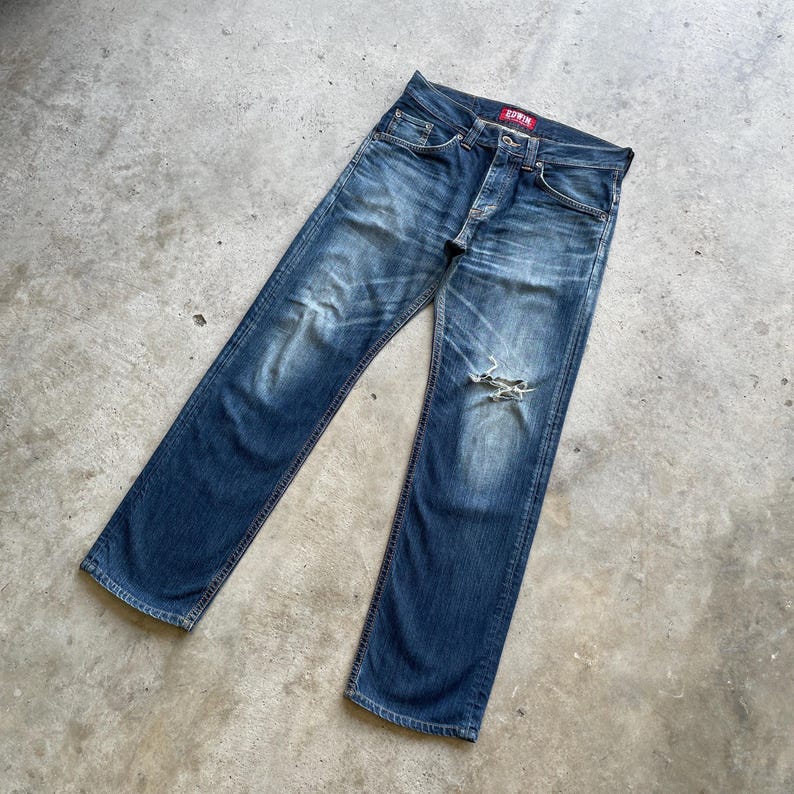 May include: Faded blue denim jeans with a distressed knee. The jeans have a button fly and a red tag on the back pocket. The jeans are laid flat on a concrete surface.