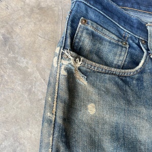 Puede incluir: Primer plano de unos vaqueros de mezclilla azul desgastados. Los vaqueros tienen un lavado descolorido, desgaste visible y un bolsillo roto. La tela muestra signos de desgaste, con roturas y parches descoloridos visibles.