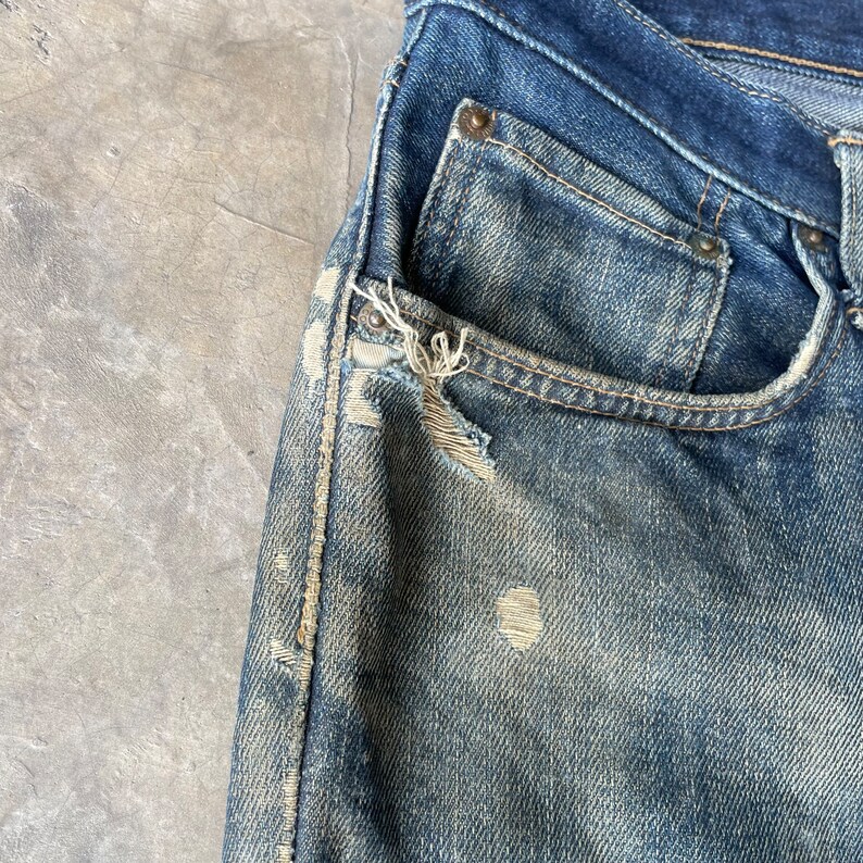 Puede incluir: Primer plano de unos vaqueros de mezclilla azul desgastados. Los vaqueros tienen un lavado descolorido, desgaste visible y un bolsillo roto. La tela muestra signos de desgaste, con roturas y parches descoloridos visibles.