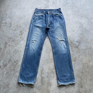 Pu&ograve; includere: Jeans in denim blu sbiadito con dettagli strappati, tra cui un ginocchio strappato e un orlo sfilacciato. I jeans hanno un classico design a cinque tasche e una chiusura con bottone e cerniera. Il denim ha un aspetto usurato.