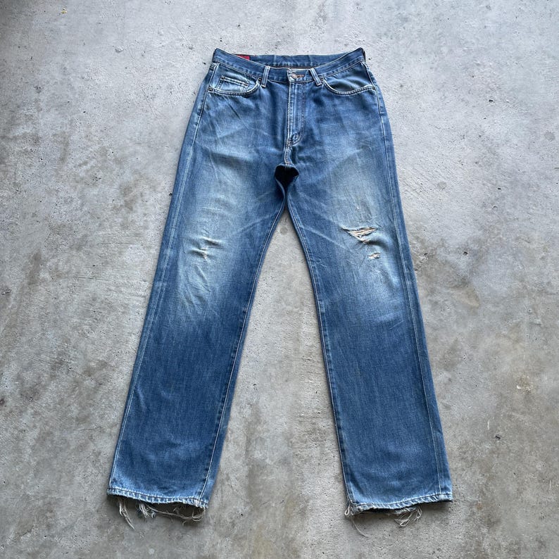 Pu&ograve; includere: Jeans in denim blu sbiadito con dettagli strappati, tra cui un ginocchio strappato e un orlo sfilacciato. I jeans hanno un classico design a cinque tasche e una chiusura con bottone e cerniera. Il denim ha un aspetto usurato.