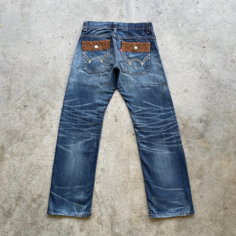 Puede incluir: Vaqueros azules con detalles de bolsillo de gamuza marr&oacute;n. Los vaqueros tienen un dise&ntilde;o cl&aacute;sico de cinco bolsillos, con el logotipo de la marca Levi's visible en el bolsillo trasero. El denim tiene un aspecto descolorido y desgastado.