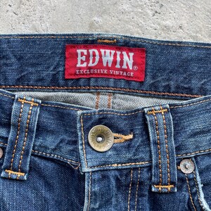 May include: Close-up of blue denim jeans with a button closure and visible stitching. A red tag with the brand name "EDWIN" and the text "EXCLUSIVE VINTAGE" is sewn on the waistband.