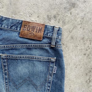 May include: Close-up of blue denim jeans with a brown leather patch displaying the brand name "EDWIN." The jeans have a classic five-pocket design, with a visible back pocket and a small label. The waist size is W34.