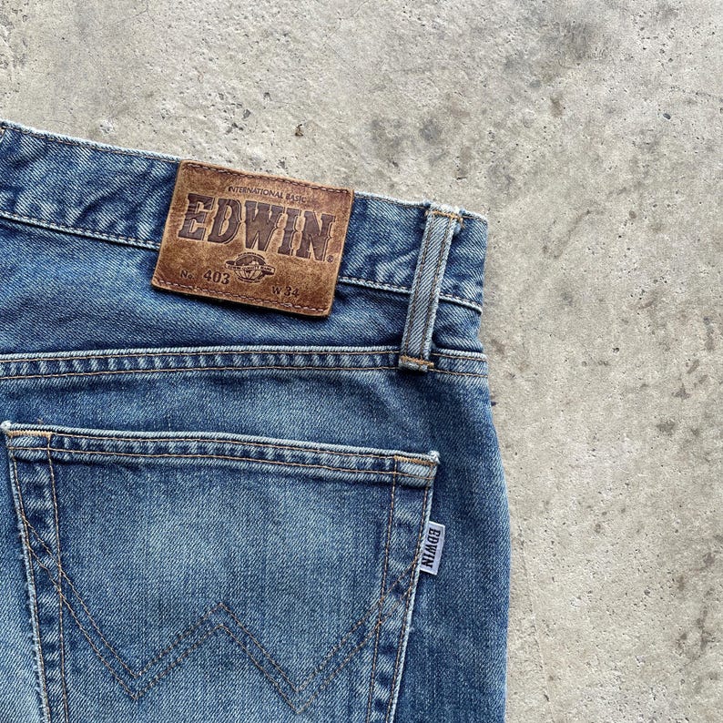 May include: Close-up of blue denim jeans with a brown leather patch displaying the brand name "EDWIN." The jeans have a classic five-pocket design, with a visible back pocket and a small label. The waist size is W34.