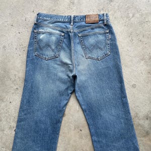 May include: Faded blue denim jeans with a classic five-pocket design. The jeans have a worn appearance with distressed areas and a leather patch at the waistband. The back pockets feature a distinctive stitched design.