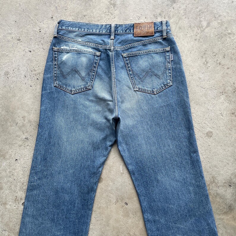 May include: Faded blue denim jeans with a classic five-pocket design. The jeans have a worn appearance with distressed areas and a leather patch at the waistband. The back pockets feature a distinctive stitched design.