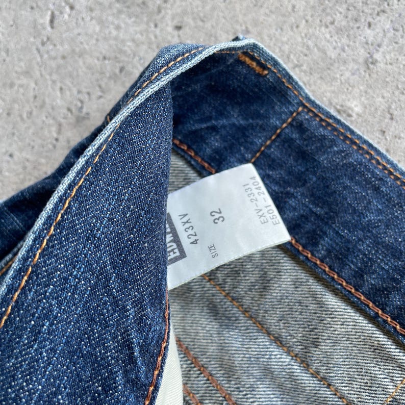 May include: Close-up of blue denim jeans with a white label. The label shows size 32 and code 423XV. The jeans feature orange stitching and a classic denim texture.