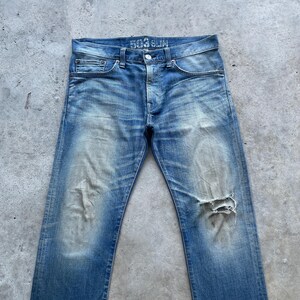 Puede incluir: Vaqueros de mezclilla azul descolorido con un aspecto desgastado. Los vaqueros tienen bragueta con botones, dise&ntilde;o cl&aacute;sico de cinco bolsillos y una rodilla rasgada. El texto "503 SLIM" es visible en la cintura.