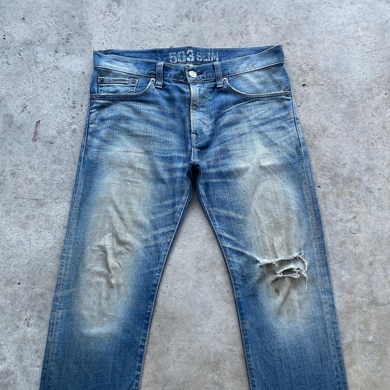 Puede incluir: Vaqueros de mezclilla azul descolorido con un aspecto desgastado. Los vaqueros tienen bragueta con botones, dise&ntilde;o cl&aacute;sico de cinco bolsillos y una rodilla rasgada. El texto "503 SLIM" es visible en la cintura.