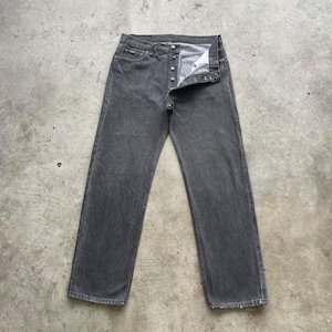 May include: A pair of gray denim jeans with a button fly. The jeans have a straight leg cut and are laid flat on a concrete surface. The denim has a faded, worn appearance.