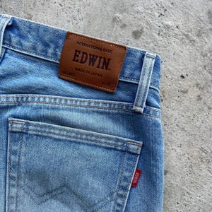 May include: Light blue denim jeans with a brown leather patch that reads "EDWIN MADE IN JAPAN". The jeans have a back pocket with a red EDWIN tag and a size 31 label.