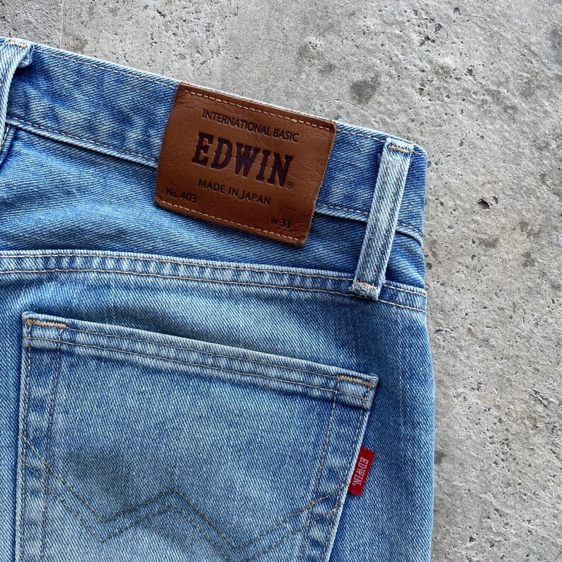 May include: Light blue denim jeans with a brown leather patch that reads "EDWIN MADE IN JAPAN". The jeans have a back pocket with a red EDWIN tag and a size 31 label.