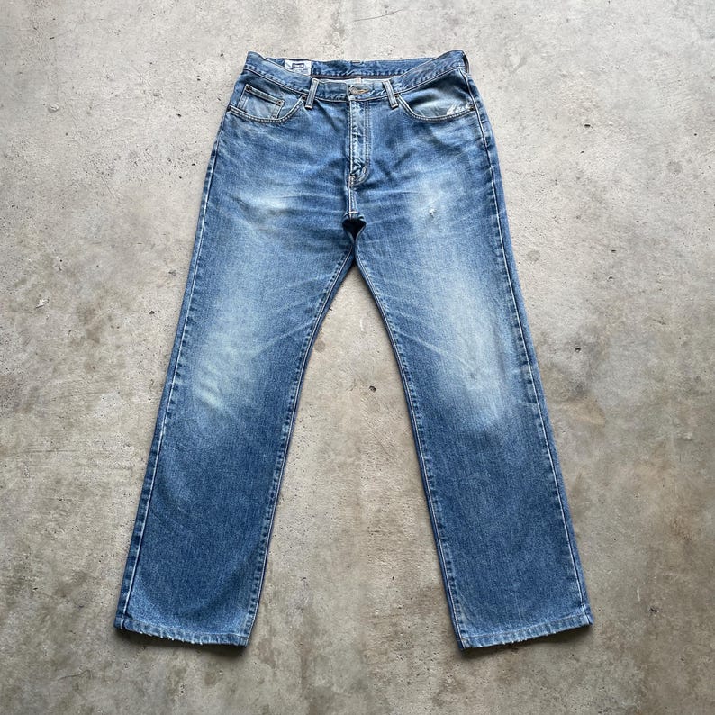 May include: Faded blue denim jeans with a classic five-pocket design. The jeans have a button fly and a straight leg cut. The denim shows signs of wear, with lighter areas on the thighs and knees. The jeans are laid flat on a concrete surface.