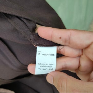May include: Close-up of a brown garment tag. The tag is white with black text that reads "NO. BI-D74-186, Fabriqu&eacute; au Japon, Made in Japan, Y's bis Co., Ltd.". The garment is a dark brown color.