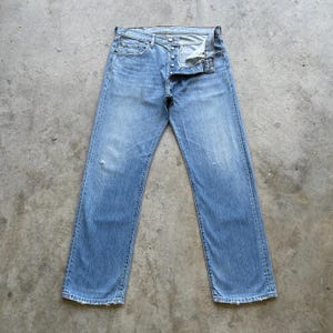 Op de afbeelding: Lichtblauwe denim jeans met een knoopsluiting. De jeans hebben een klassiek vijf-zakken ontwerp en een licht versleten look met een kleine scheur op het linkerbeen. De jeans liggen plat op een betonnen oppervlak.