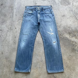 May include: Light blue denim jeans with a classic five-pocket design. The jeans have a faded wash and distressed details, including rips on the knees. The jeans are laid flat on a concrete surface.