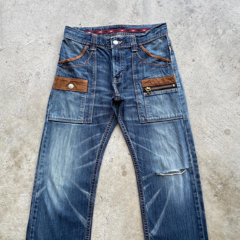 Puede incluir: Vaqueros de mezclilla azules con detalles de bolsillo &uacute;nicos. Incluyen detalles en marr&oacute;n tipo ante, bolsillos con botones y cremalleras, y una rodilla desgastada. Los vaqueros tienen un dise&ntilde;o cl&aacute;sico de cinco bolsillos y bragueta con botones.