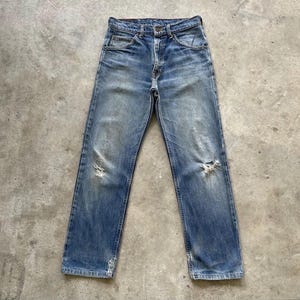 May include: Faded blue denim jeans with distressed details, including rips at the knees. The jeans have a classic five-pocket design, a button and zipper closure, and a worn appearance.