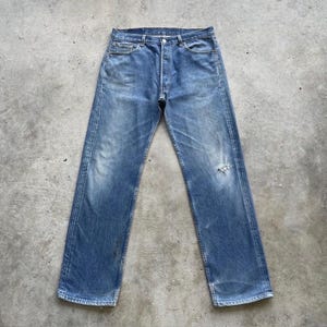 May include: Faded blue denim jeans with a button fly and a distressed knee. The jeans have a classic five-pocket design and a straight leg fit. The denim has a worn appearance.