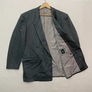 Può includere: Un blazer grigio a doppio petto con un sottile motivo a righe verticali. Il blazer ha un revers a lancia, due tasche anteriori e una fodera interna grigio chiaro. All'interno è visibile un'etichetta con la scritta "Christian Dior".