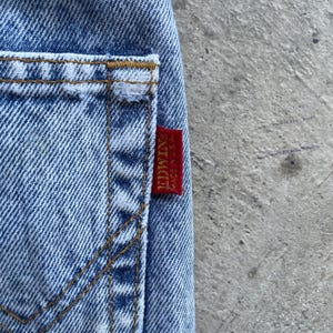May include: Close-up of light blue denim jeans with a visible pocket and stitching. A red tag with the brand name "EDWIN" and "MADE IN U.S.A." is attached to the jeans. The jeans have a classic, worn look.