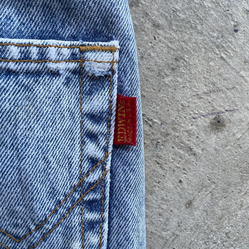 May include: Close-up of light blue denim jeans with a visible pocket and stitching. A red tag with the brand name "EDWIN" and "MADE IN U.S.A." is attached to the jeans. The jeans have a classic, worn look.