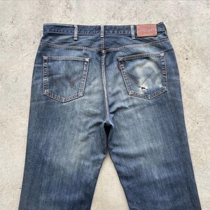K&ouml;nnte beinhalten: Verblasste blaue Jeans mit klassischem F&uuml;nf-Taschen-Design. Die R&uuml;ckansicht zeigt die Taschen, G&uuml;rtelschlaufen und einen Lederaufn&auml;her. Die Jeans haben ein abgenutztes Aussehen mit einem kleinen Riss an einer der Ges&auml;&szlig;taschen.