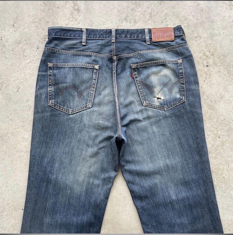 K&ouml;nnte beinhalten: Verblasste blaue Jeans mit klassischem F&uuml;nf-Taschen-Design. Die R&uuml;ckansicht zeigt die Taschen, G&uuml;rtelschlaufen und einen Lederaufn&auml;her. Die Jeans haben ein abgenutztes Aussehen mit einem kleinen Riss an einer der Ges&auml;&szlig;taschen.