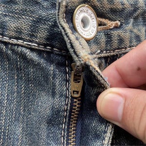May include: Close-up of blue denim jeans with a brass zipper and a button. The button is white with the word "Wrangler" in a circular design. The zipper is partially open, revealing the denim's texture.