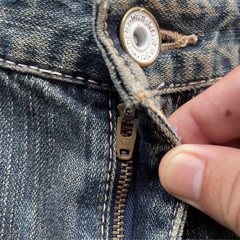 May include: Close-up of blue denim jeans with a brass zipper and a button. The button is white with the word "Wrangler" in a circular design. The zipper is partially open, revealing the denim's texture.