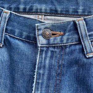Pu&ograve; includere: Primo piano di jeans di denim blu. I jeans presentano un bottone metallico con inciso il nome del marchio "EDWIN". Le cuciture sono visibili e il tessuto ha un aspetto usurato.