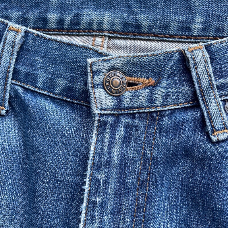 Pu&ograve; includere: Primo piano di jeans di denim blu. I jeans presentano un bottone metallico con inciso il nome del marchio "EDWIN". Le cuciture sono visibili e il tessuto ha un aspetto usurato.